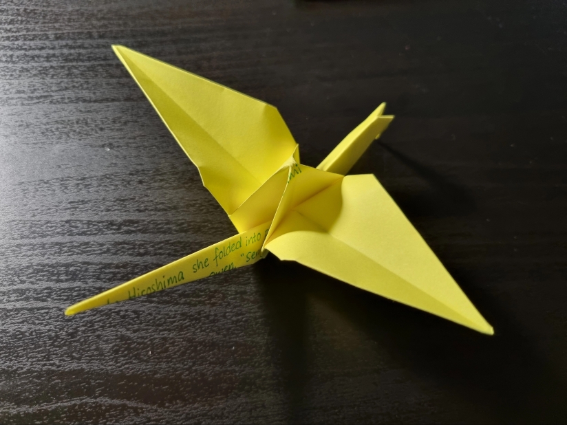 Yellow paper origami crane on a black table. On the paper, in green pen, are written fragments of a poem not fully visible.