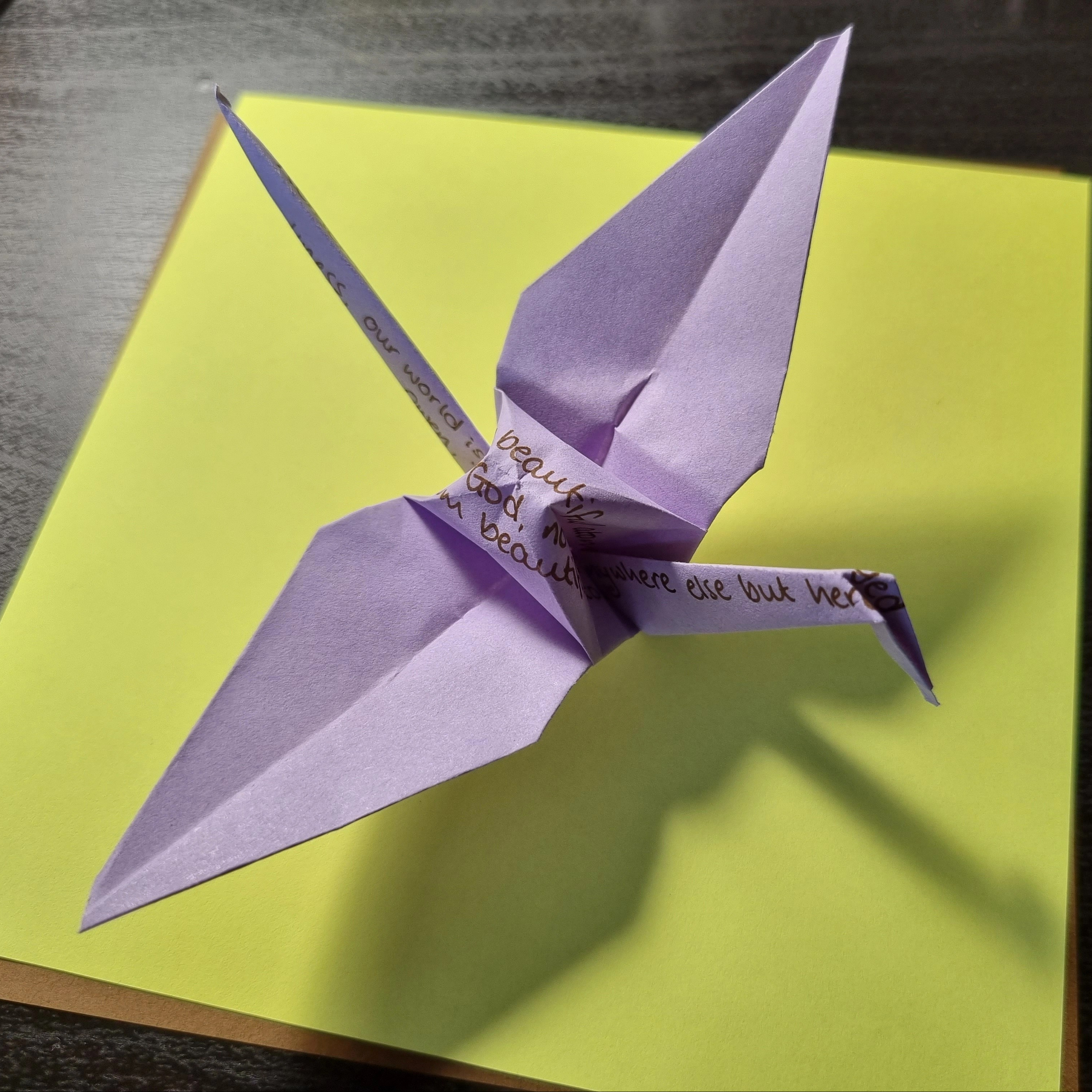 Lilac paper origami crane with fragments of a poem in brown pen, placed on yellow square paper on black table.