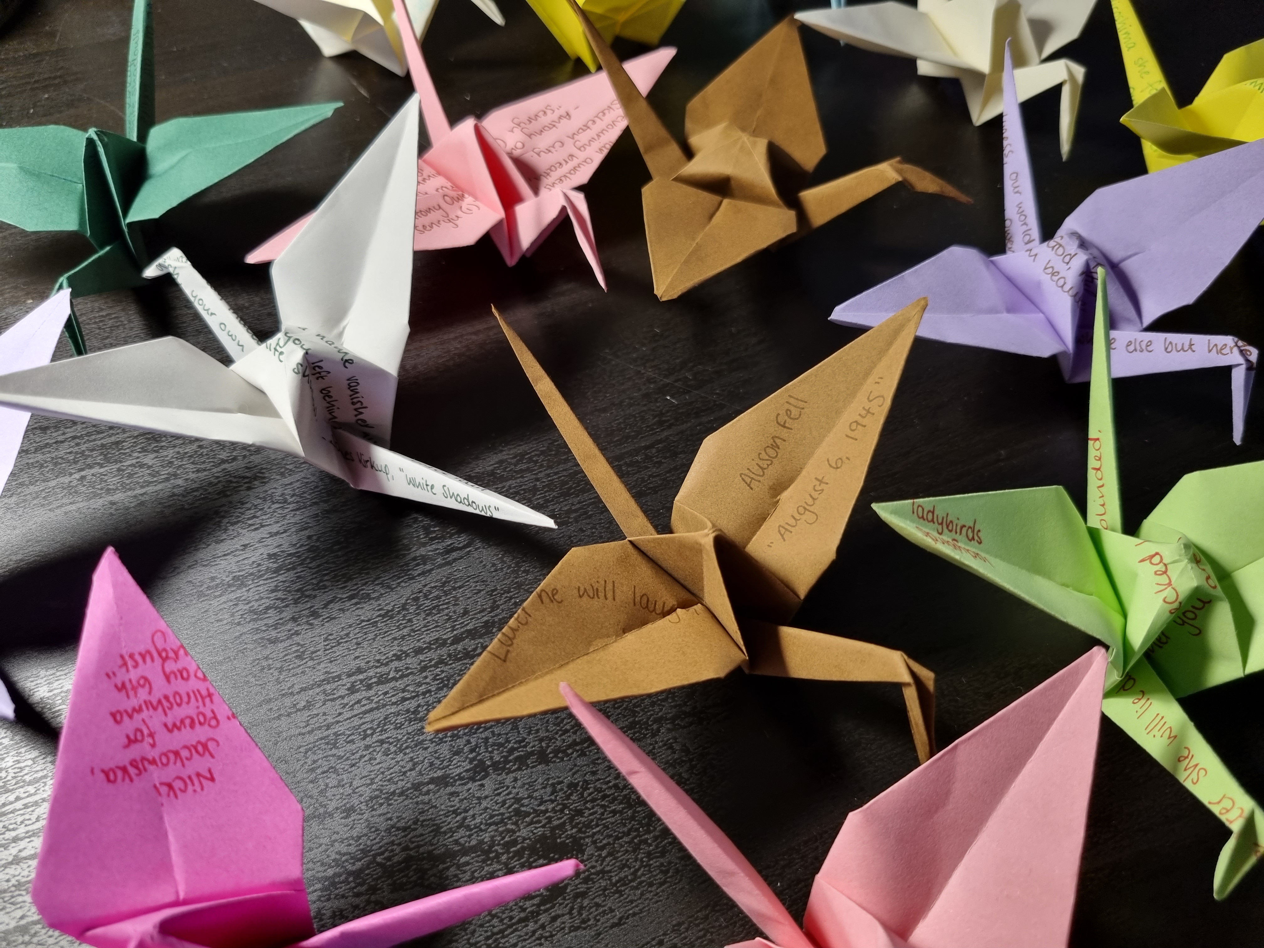 Colourful origami paper cranes on a black table. Some have fragments of poems written on them.
