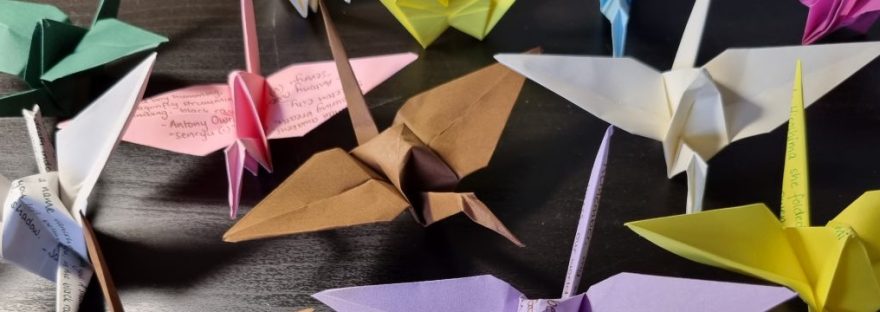 Colourful origami paper cranes on a black table. Some have fragments of poems written on the folded paper.