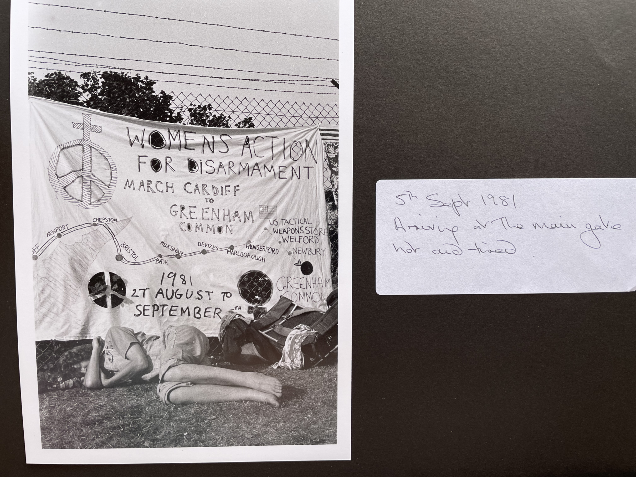 Scrapbook page. On the left, a black and white photograph in portrait orientation. A woman, arm across her face, lies outside on the grassy ground at the foot of a banner hung on a chain link fence. A large rucksack is beside her. The hand-drawn banner reads 'Women's action for disarmament march Cardiff to Greenham Common' and has the dates '1981 27th August to September 5th'. A map of the route is also drawn on the banner. To the right of the photo, a white sticker has the hand written caption: '5th Sept 1981. Arriving at the main gate hot and tired'.