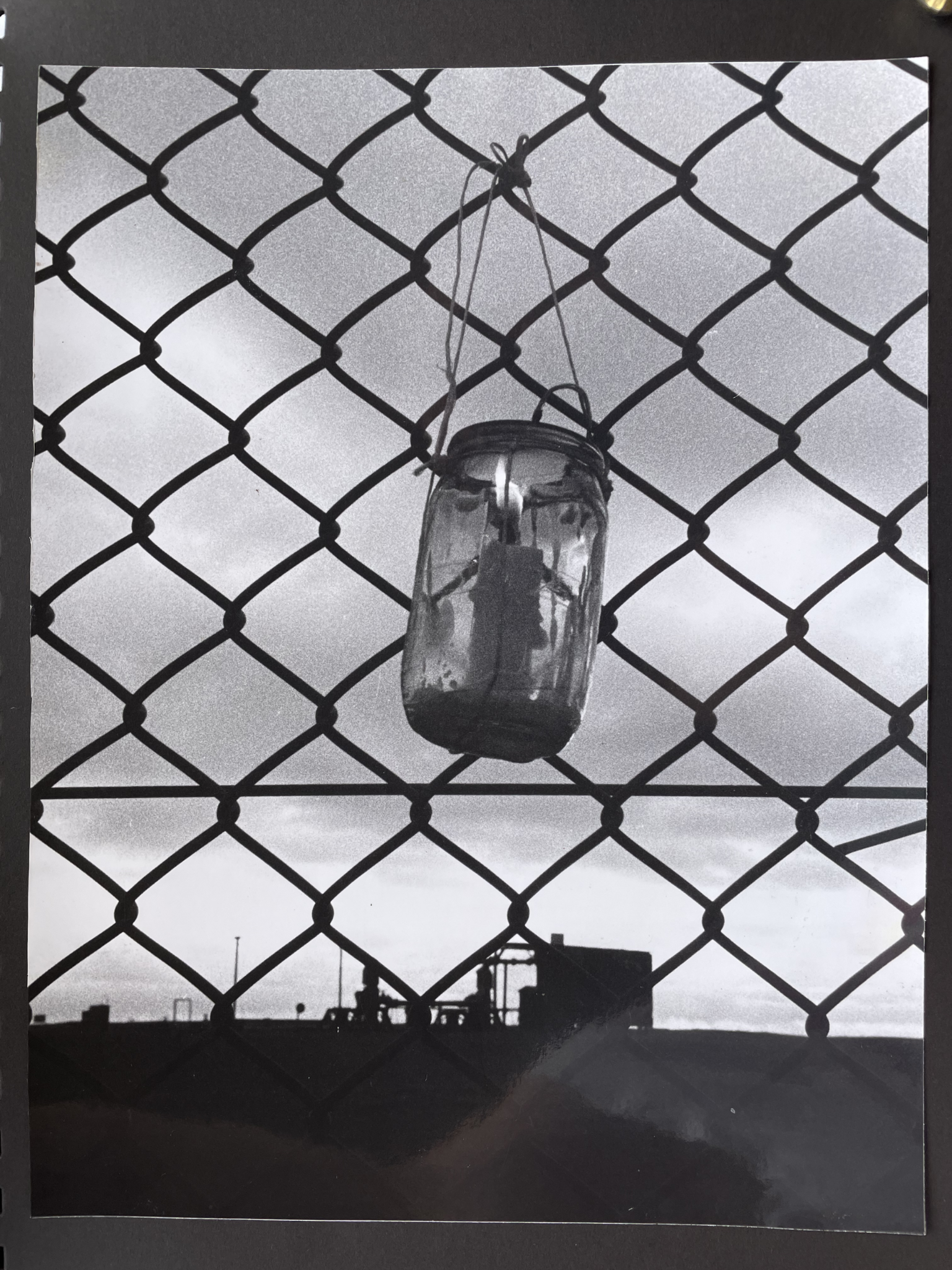 Scrapbook page with black and white photo in portrait orientation. Photo shows a jar containing a burning candle. The jar hangs from string tied to a chain-link fence. Through the fence, the view is mostly sky, with the shape of military buildings seen as silhouettes at the bottom of the image.