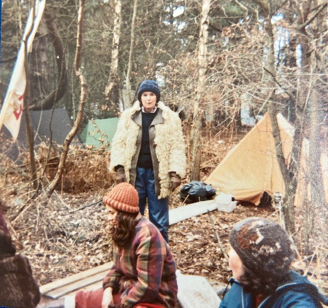 Photograph shows three women in the woods at the Greenham Common Women's Peace Camp. A woman in a pale fluffy coat, blue jeans, and blue knitted hat stands centrally and looks directly facing the camera. The other women, also wearing hats and coats, sit in front of her, looking away. In the background there are tents. A banner hangs from a tree. Trees have bare branches, and the ground is covered with brown leaves.