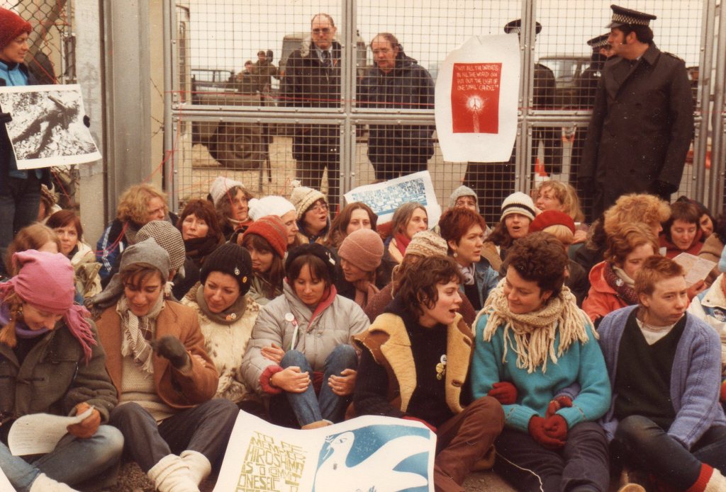 Colour photograph. Outside. In the lower half of the image, a group of around 30 women, many in coats, cardigans, scarves and gloves, sit on the ground. Behind their backs is a wire gate, seen in the top half of the image. Men in dark coats look through the wire at the women. One policeman stands in front of the wire, between the women and the gate, speaking to two policemen behind the gate. The women display posters. On the gate, a red poster has a slogan above an image of a candle with a CND symbol in the flame. A woman stands on the left of the photo holding a large black and white image of a nuclear bombing victim. On the ground in front of the women is a op poster with an image of a dove, and text mentioning Hiroshima.